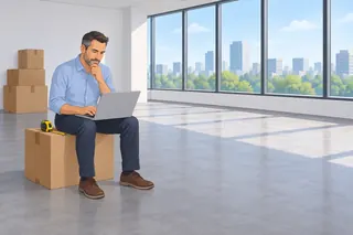 An office manager sits on a moving box in an empty new office with a laptop and tape measure. There are no desks yet.
